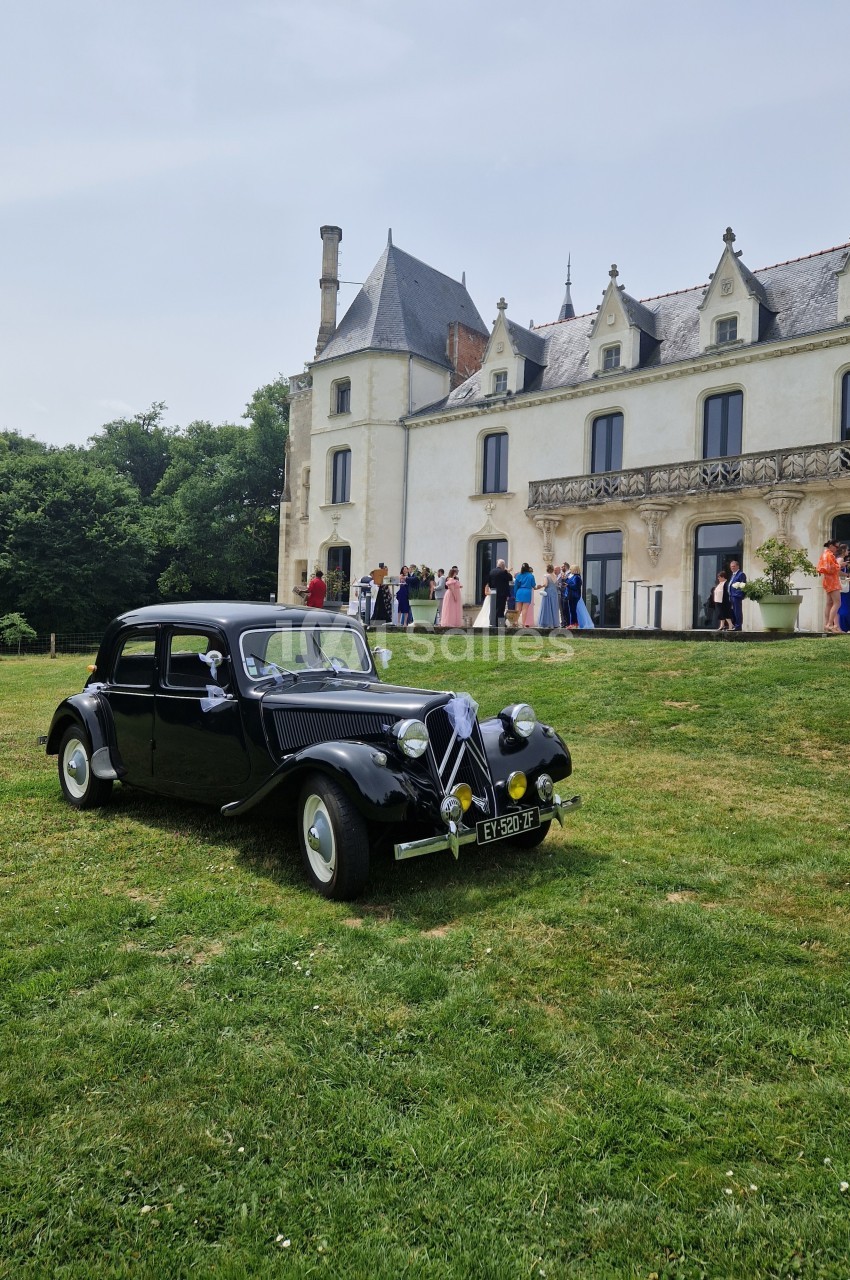 Voiture ancienne noire garée sur une pelouse devant un château, avec des invités en tenue élégante à l'arrière-plan.