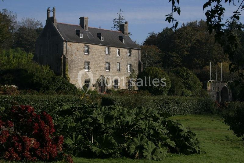 Manoir en pierre entouré de jardins verdoyants et d'arbres, sous un ciel légèrement voilé.