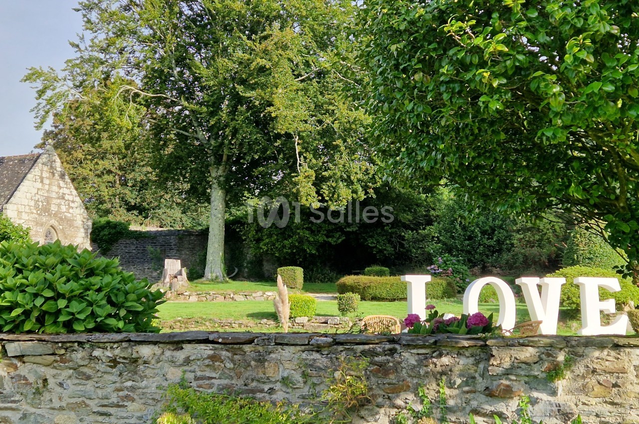 Jardin verdoyant avec des buissons taillés, une église en pierre et des lettres blanches formant ’LOVE’ sur un muret.