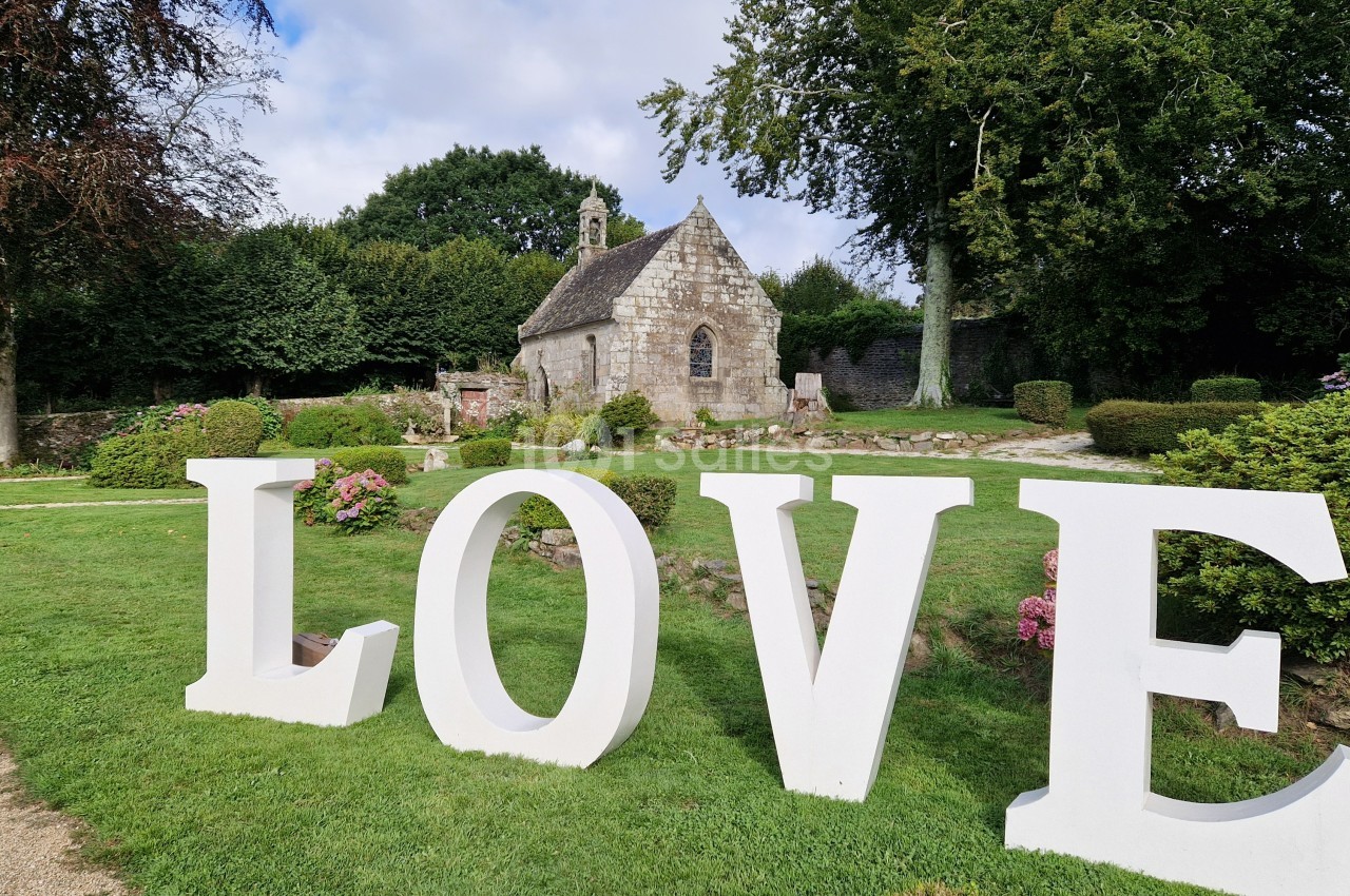 Grandes lettres blanches formant le mot ’LOVE’ sur une pelouse devant une petite chapelle en pierre entourée de verdure.