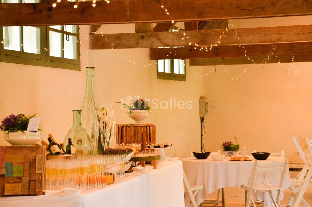 Table de réception décorée avec des boissons, des fleurs et des plats, dans une salle lumineuse avec poutres apparentes.