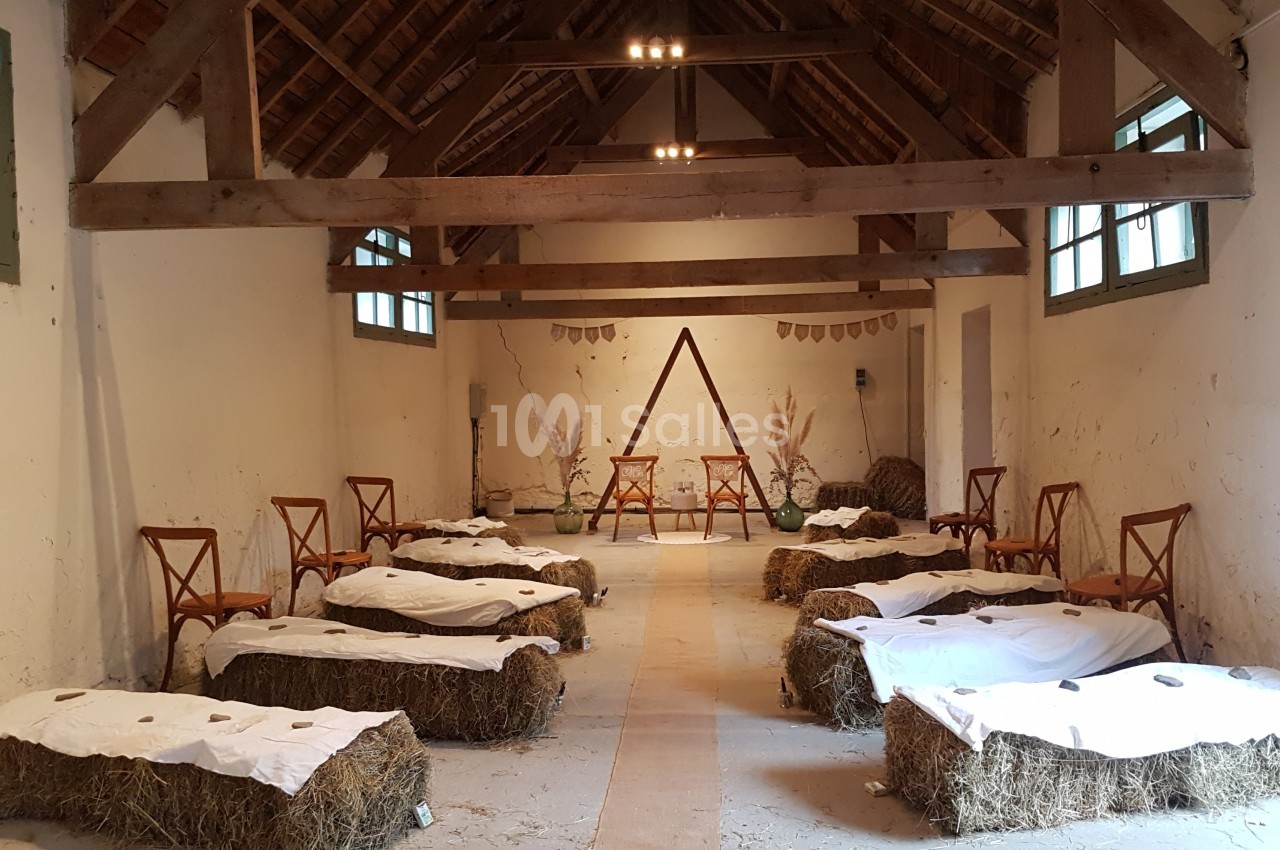 Salle rustique aménagée pour un événement, avec des bottes de paille recouvertes de tissu blanc et des chaises en bois.