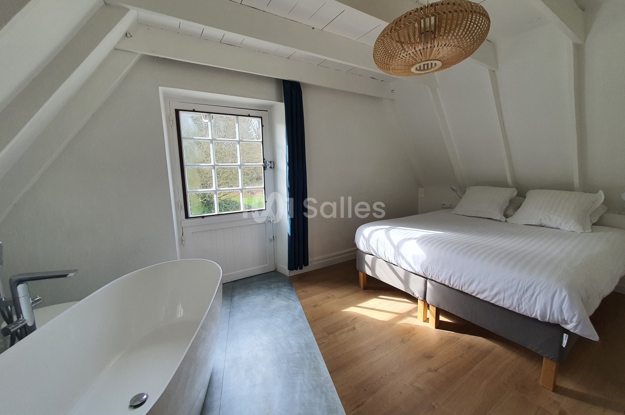 Chambre lumineuse avec lit double, baignoire blanche, parquet en bois et fenêtre donnant sur un paysage verdoyant.