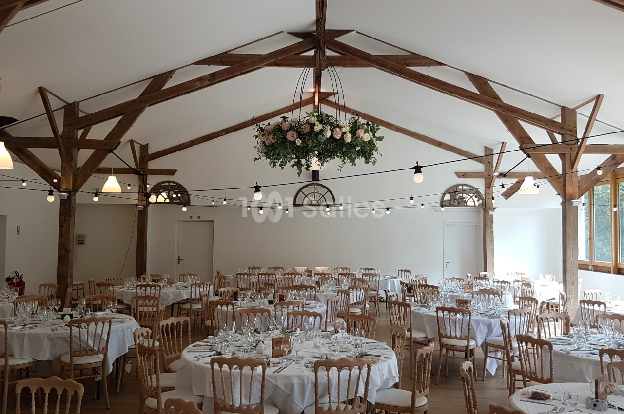 Salle de réception lumineuse avec tables rondes dressées, chaises en bois et décoration florale suspendue.