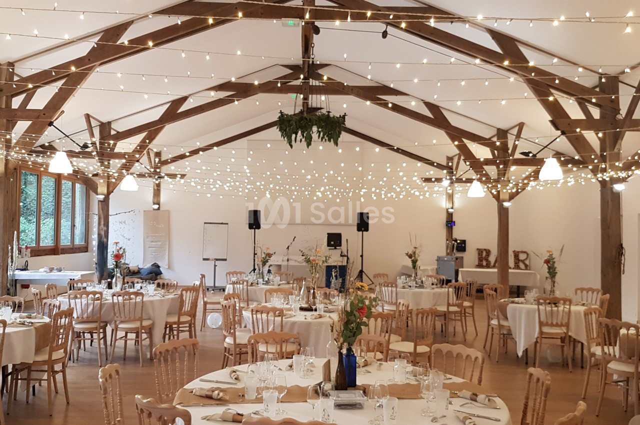 Salle de réception décorée avec des guirlandes lumineuses, des tables rondes dressées et des chaises en bois clair.