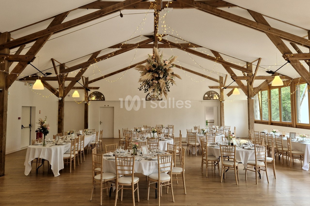 Salle de réception lumineuse avec poutres apparentes, tables dressées et décoration florale suspendue.