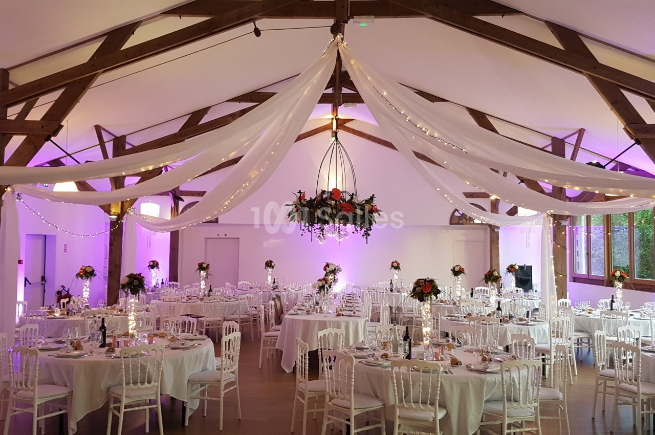 Salle de réception décorée avec des tables rondes, des chaises blanches, des drapés suspendus et un éclairage chaleureux.