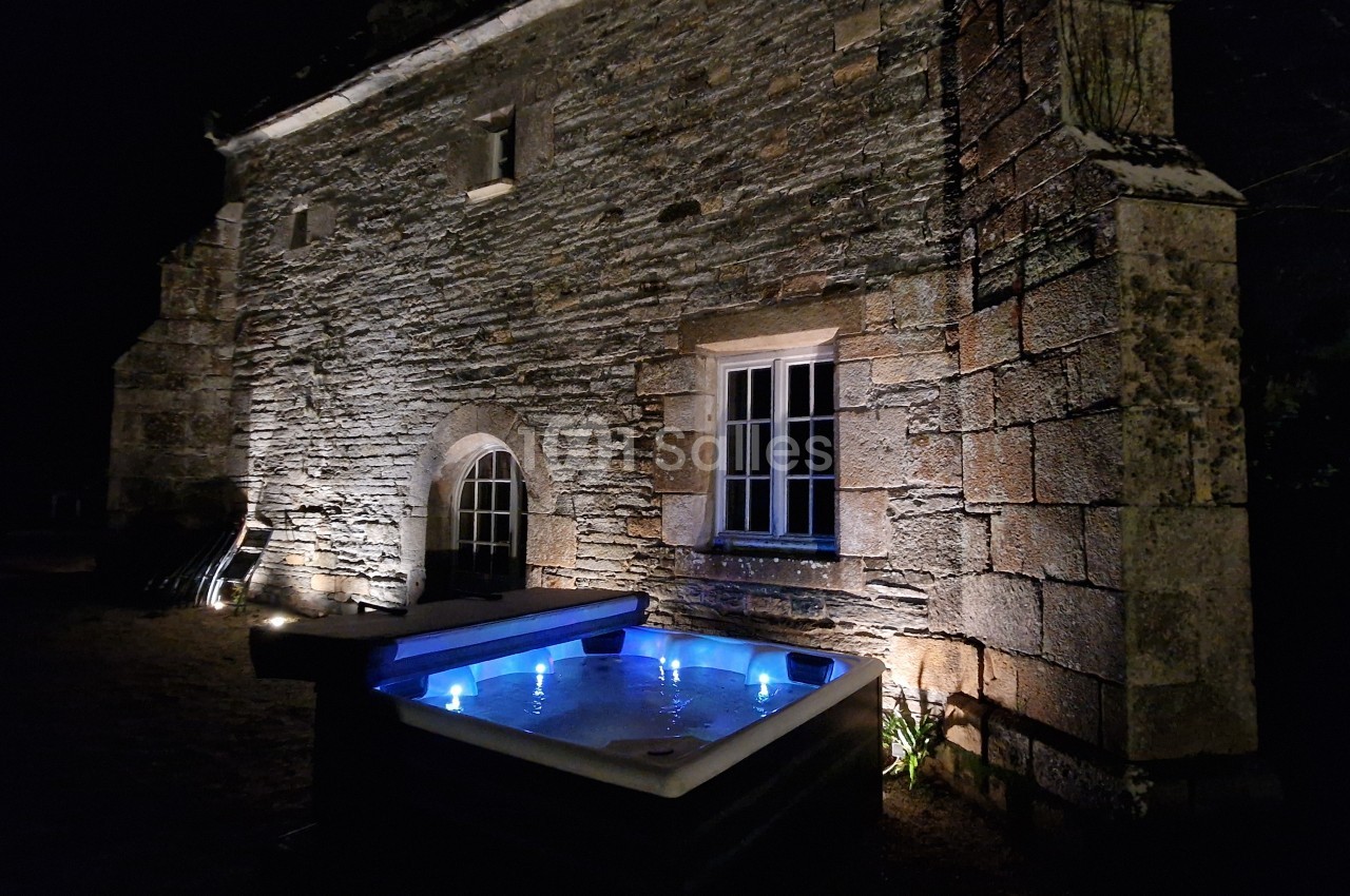 Façade en pierre d'une maison ancienne éclairée la nuit, avec un jacuzzi illuminé au premier plan.