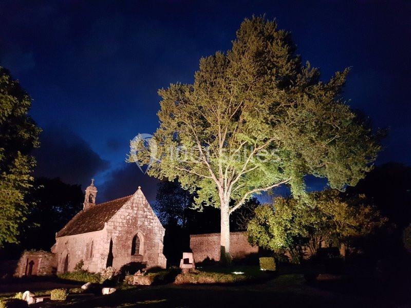 Église en pierre éclairée de nuit, entourée d'arbres et d'un jardin sous un ciel sombre.