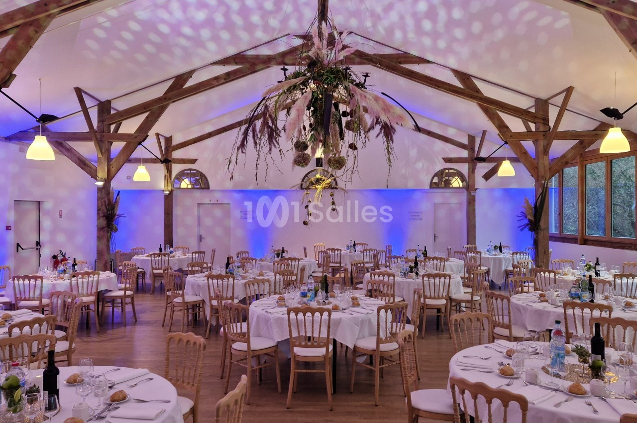 Salle de réception décorée avec des tables rondes dressées, un éclairage tamisé et une suspension florale centrale.