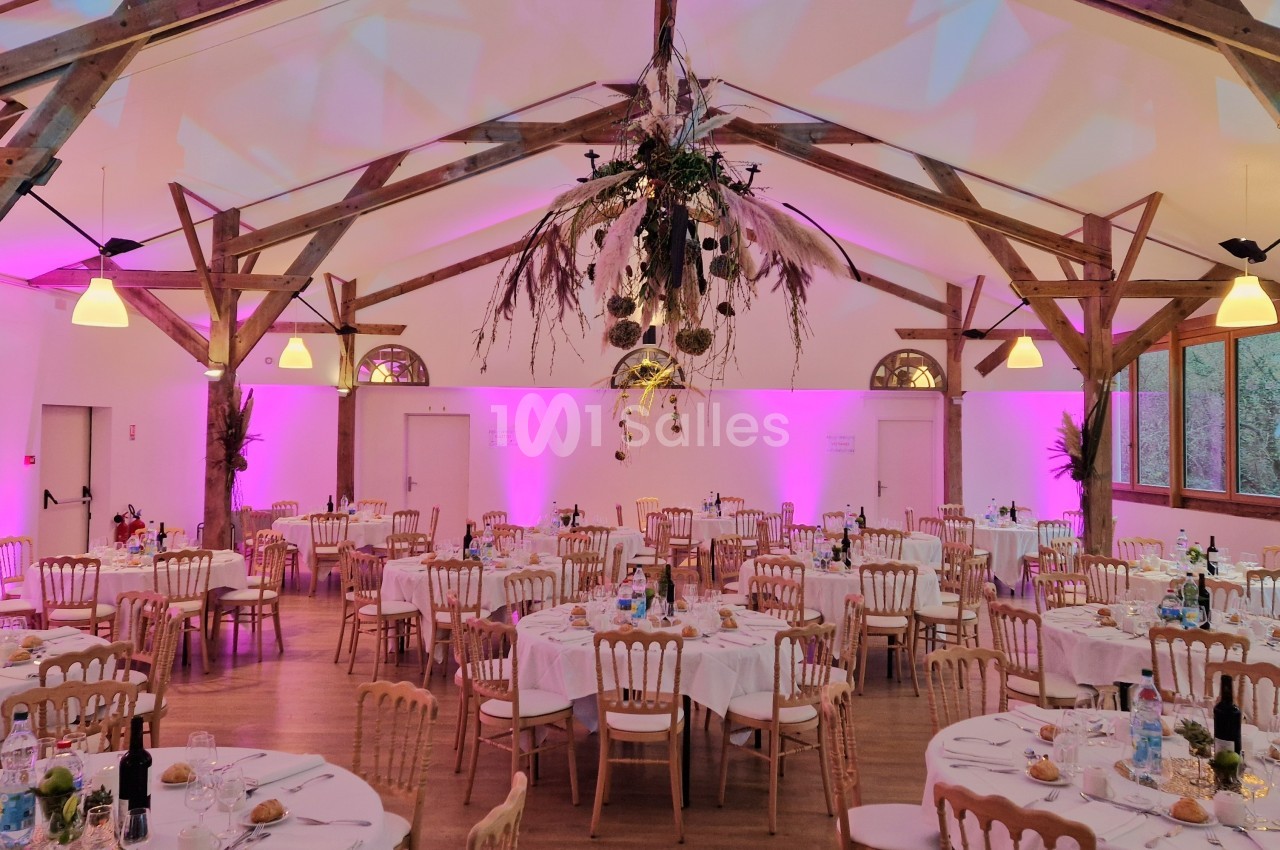 Salle de réception décorée avec des tables rondes dressées, éclairage rose et suspension florale centrale.