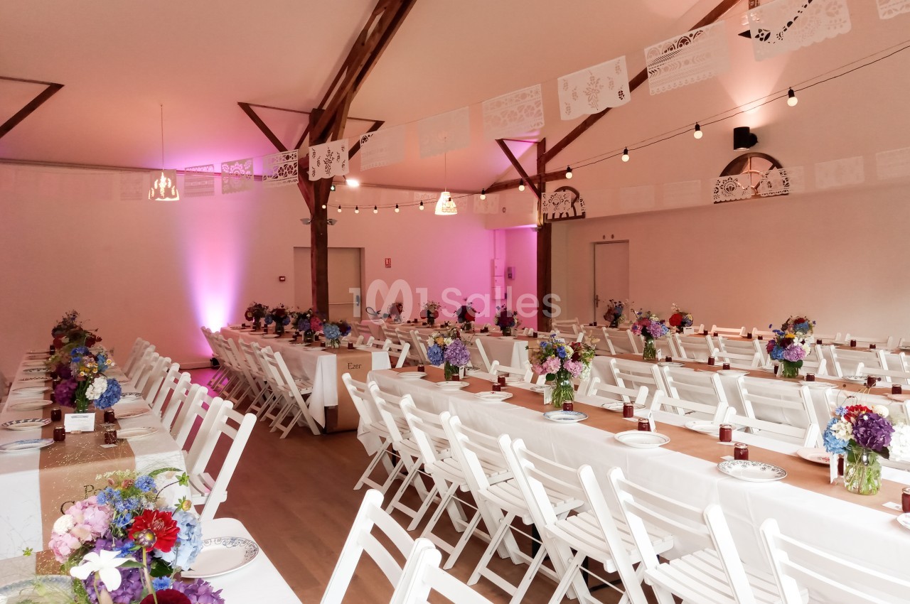Salle décorée pour un événement, avec longues tables blanches, chaises pliantes, fleurs colorées et guirlandes suspendues.
