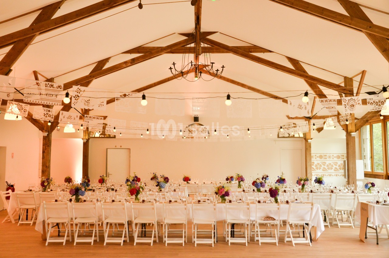Salle de réception décorée avec des guirlandes en papier, des tables alignées et des bouquets de fleurs colorées.