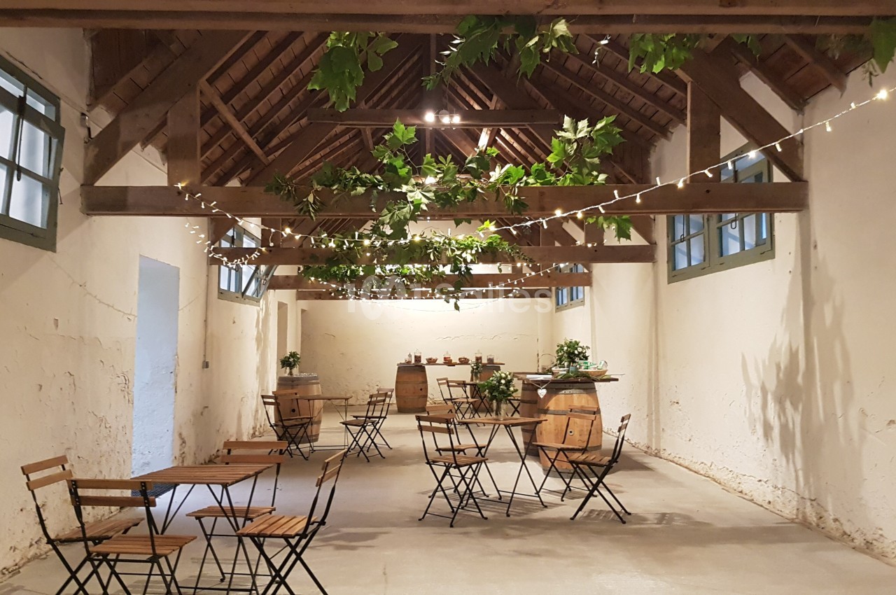 Salle rustique avec charpente en bois, tables et chaises pliantes, décorée de guirlandes lumineuses et feuillages suspendus.