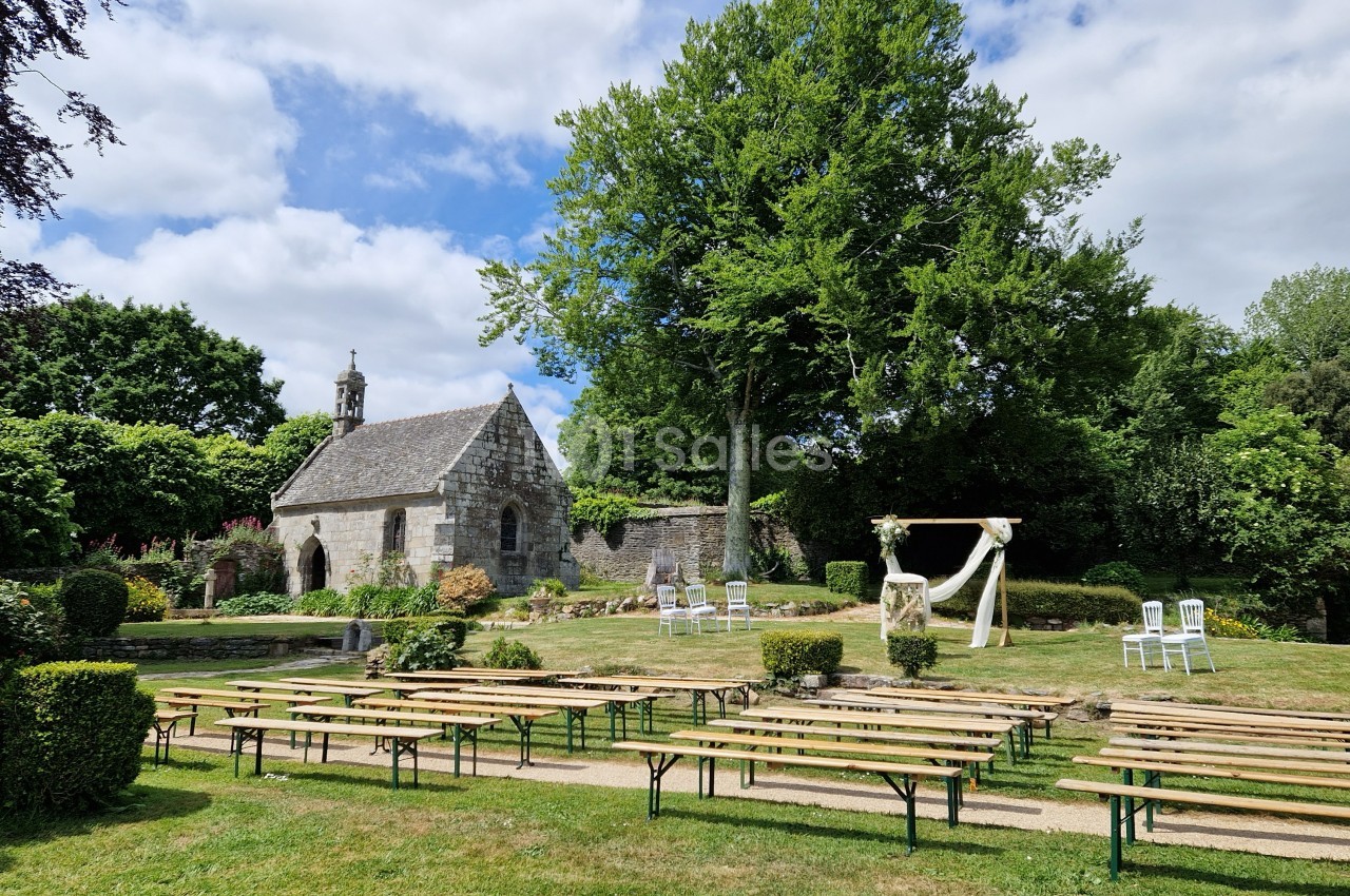 Chapelle en pierre entourée de verdure avec des bancs en bois disposés pour un événement en plein air.