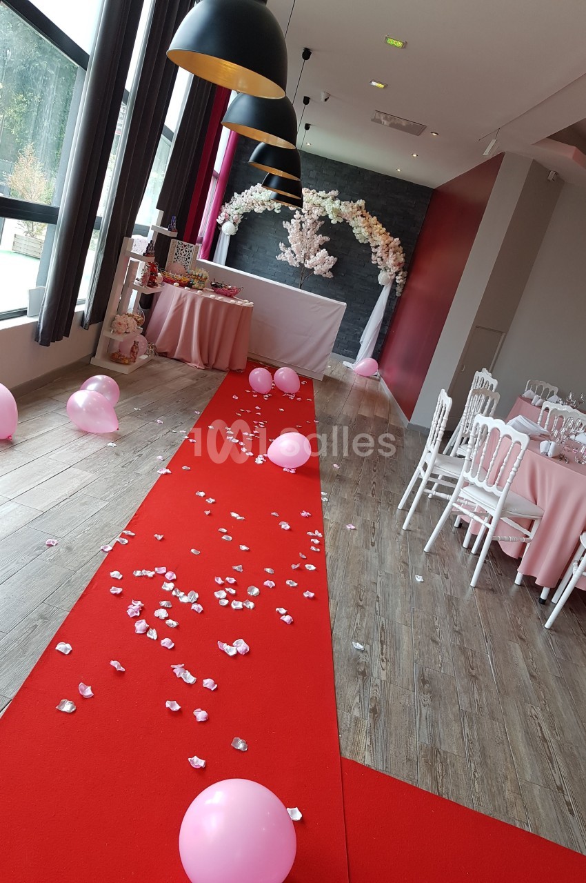 Salle décorée pour un événement avec tapis rouge, pétales, ballons roses et arche florale blanche en arrière-plan.