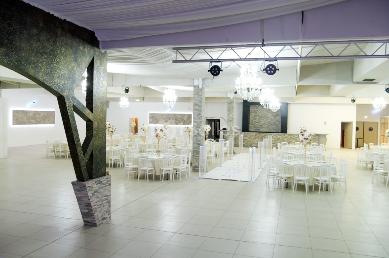 Salle de réception spacieuse avec tables rondes dressées, décorations florales et éclairage élégant.