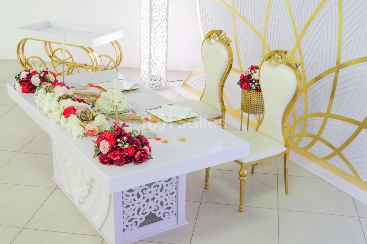 Table décorée avec des fleurs rouges et blanches, deux chaises dorées et un fond orné de motifs géométriques.