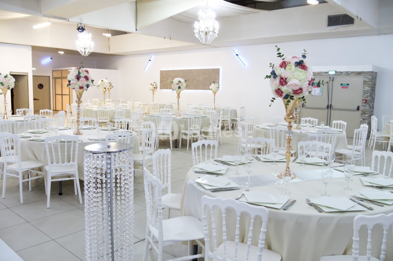 Salle de réception décorée avec des tables rondes, nappes blanches, chaises assorties et centres de table fleuris.