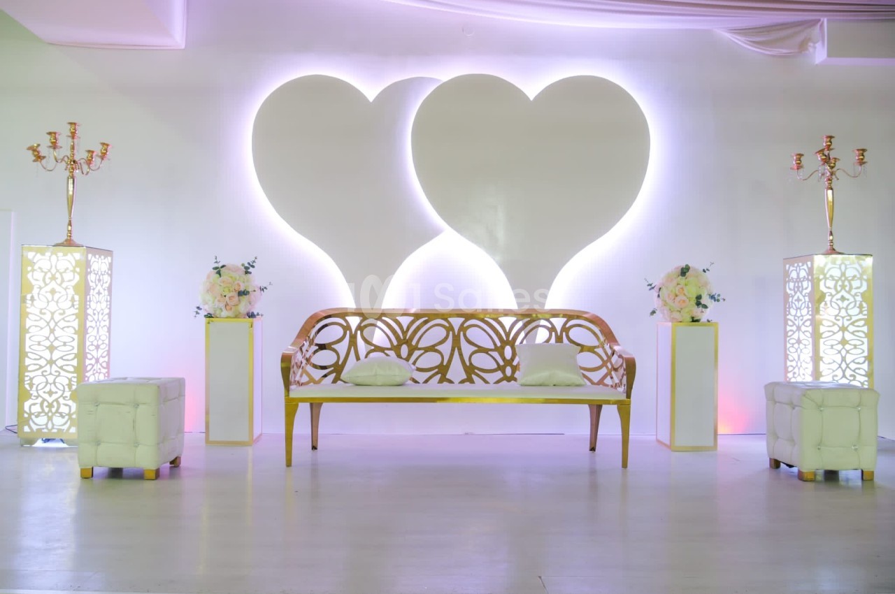 Banc décoratif doré devant un mur blanc orné de deux cœurs lumineux, entouré de bougeoirs et de fleurs.