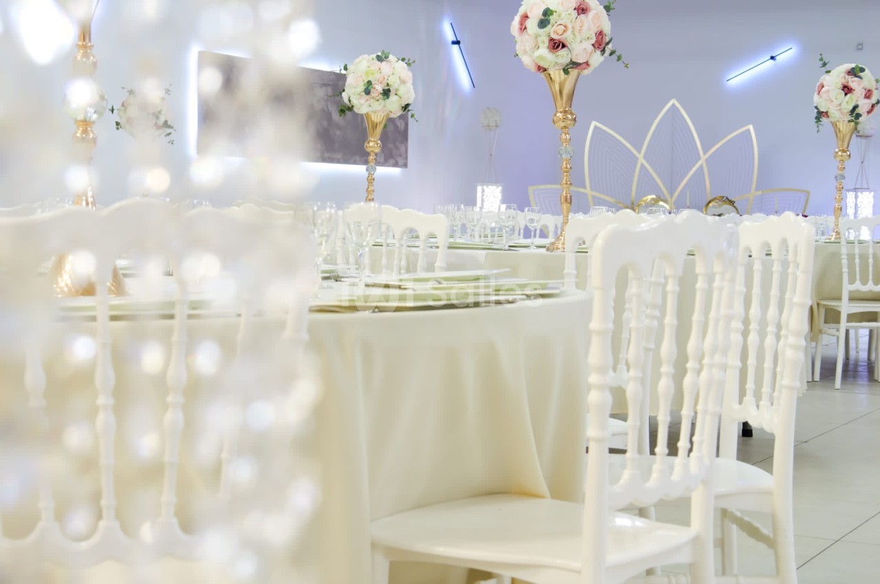 Salle de réception décorée avec des tables rondes, nappes blanches, chaises assorties et centres de table fleuris.
