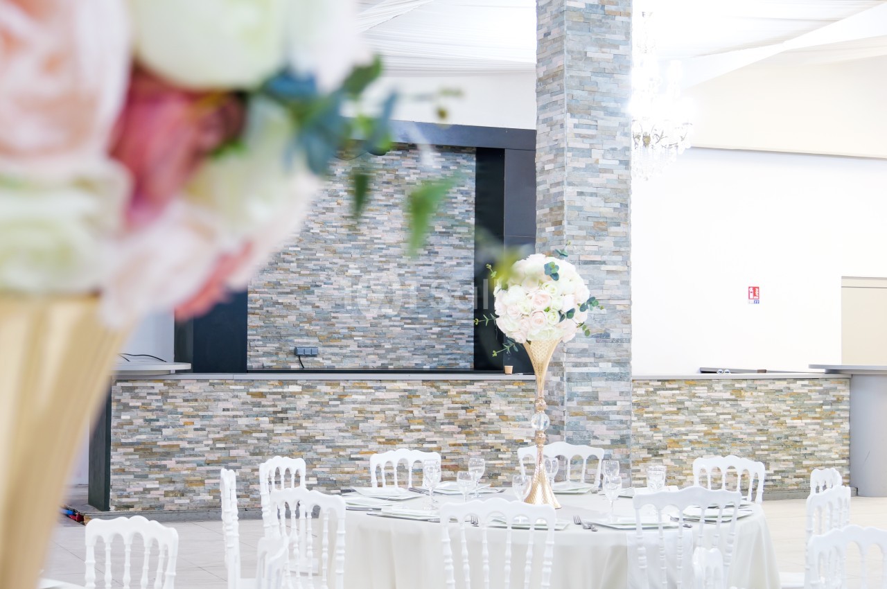 Salle de réception lumineuse avec tables décorées de centres de table fleuris et un mur en pierre en arrière-plan.