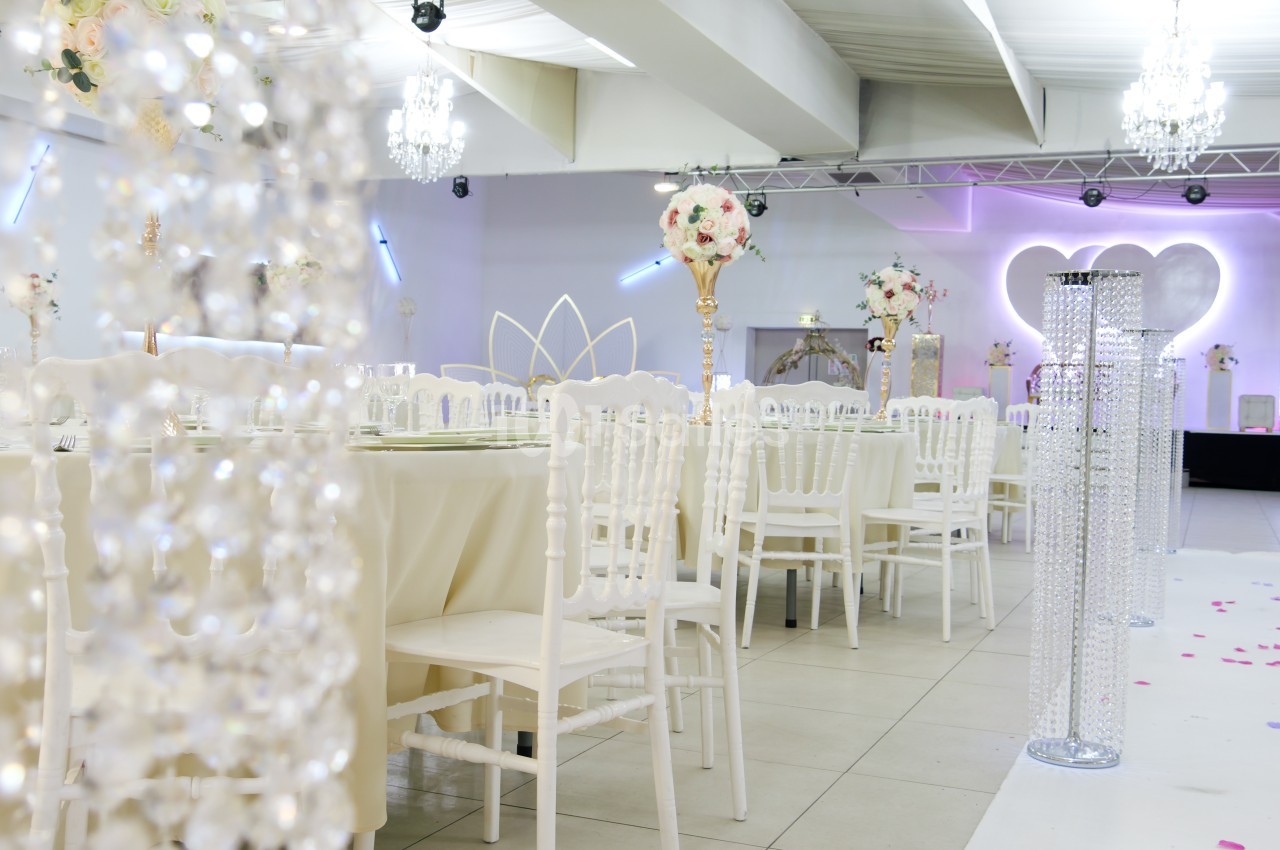 Salle de réception décorée pour un mariage avec des tables dressées, des chaises blanches et des centres de table fleuris.