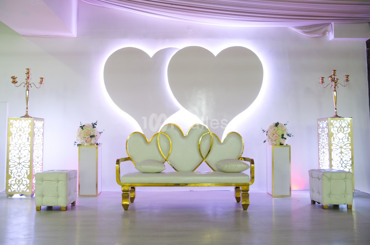 Canapé blanc et doré avec motifs de cœurs, entouré de décorations florales et éclairages doux dans une salle.