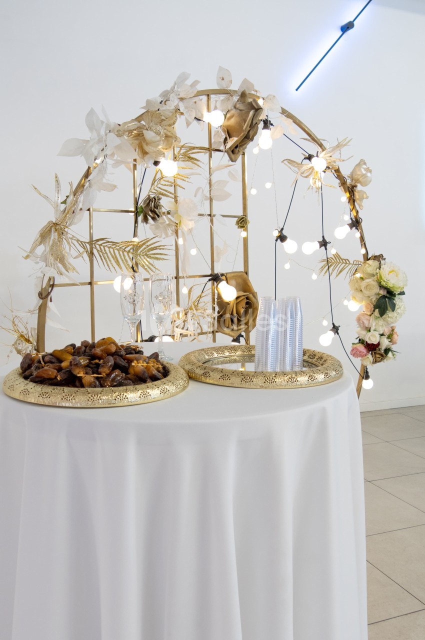 Décoration de table avec structure dorée ornée de fleurs, guirlandes lumineuses et plateaux contenant des dattes.