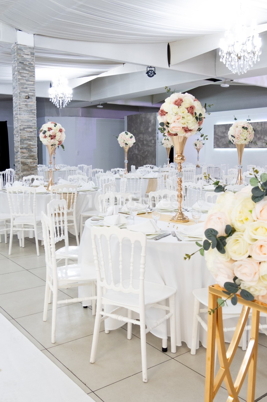 Salle de réception décorée avec des tables rondes, nappes blanches, chaises blanches et centres de table fleuris.