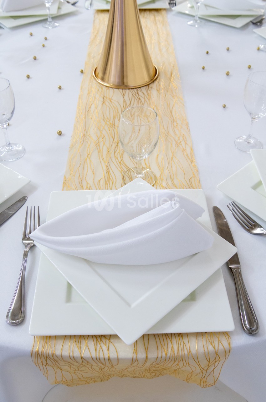 Table dressée avec une nappe blanche, un chemin de table doré, des assiettes carrées et des serviettes pliées.