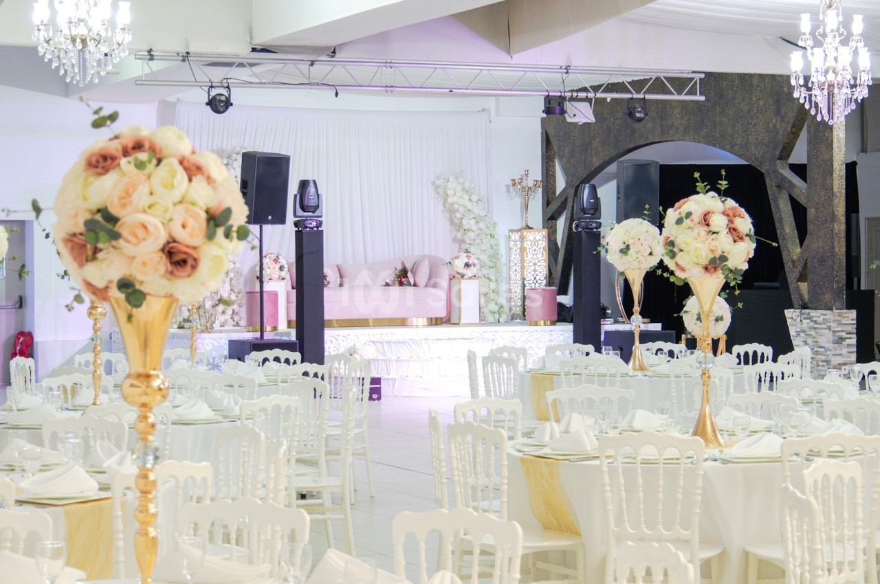 Salle de réception élégante décorée de tables rondes, chaises blanches et centres de table fleuris dans des tons pastel.