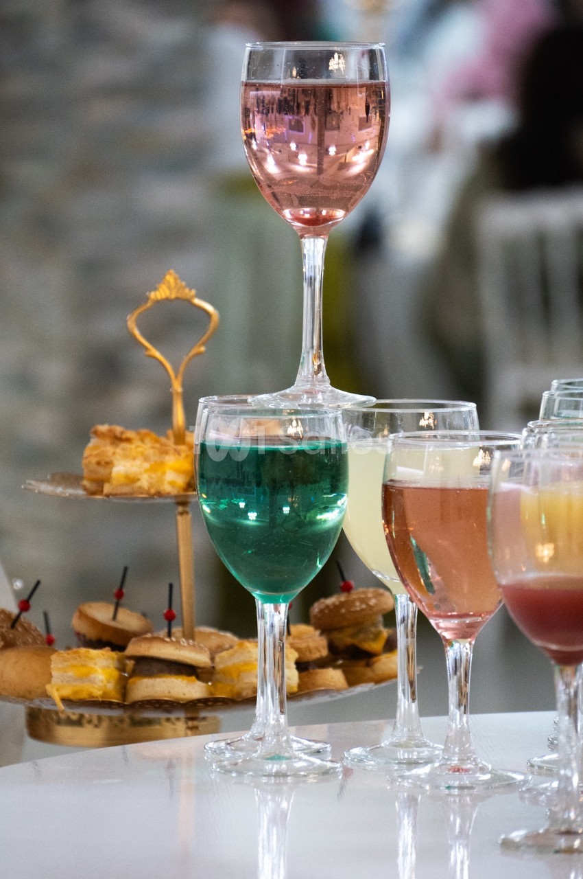 Verres de cocktails colorés sur une table, accompagnés d'un plateau de petits fours en arrière-plan flou.