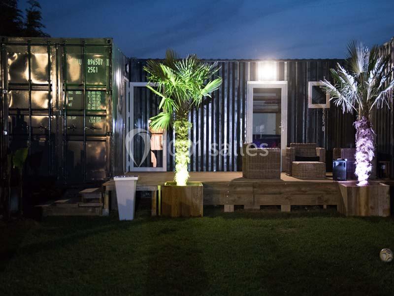 Un container aménagé en espace de vie avec terrasse éclairée, palmiers et mobilier extérieur, de nuit.