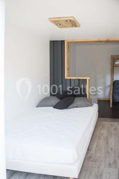 Lit double avec drap blanc et oreillers gris dans une pièce lumineuse au sol en bois, avec cloison en bois et métal.