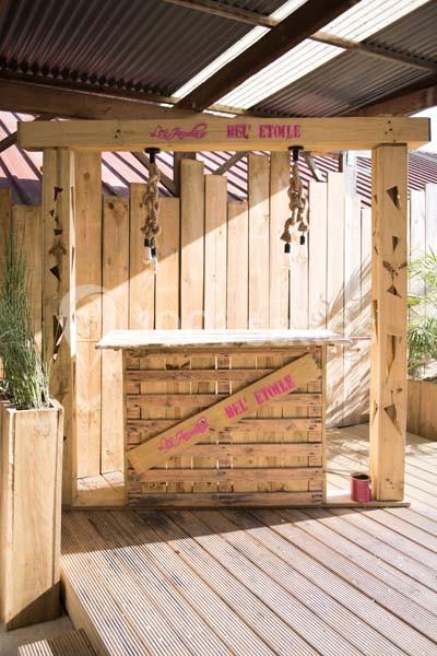 Structure en bois avec un comptoir et des ampoules suspendues, située sur une terrasse ensoleillée avec un toit en tôle.