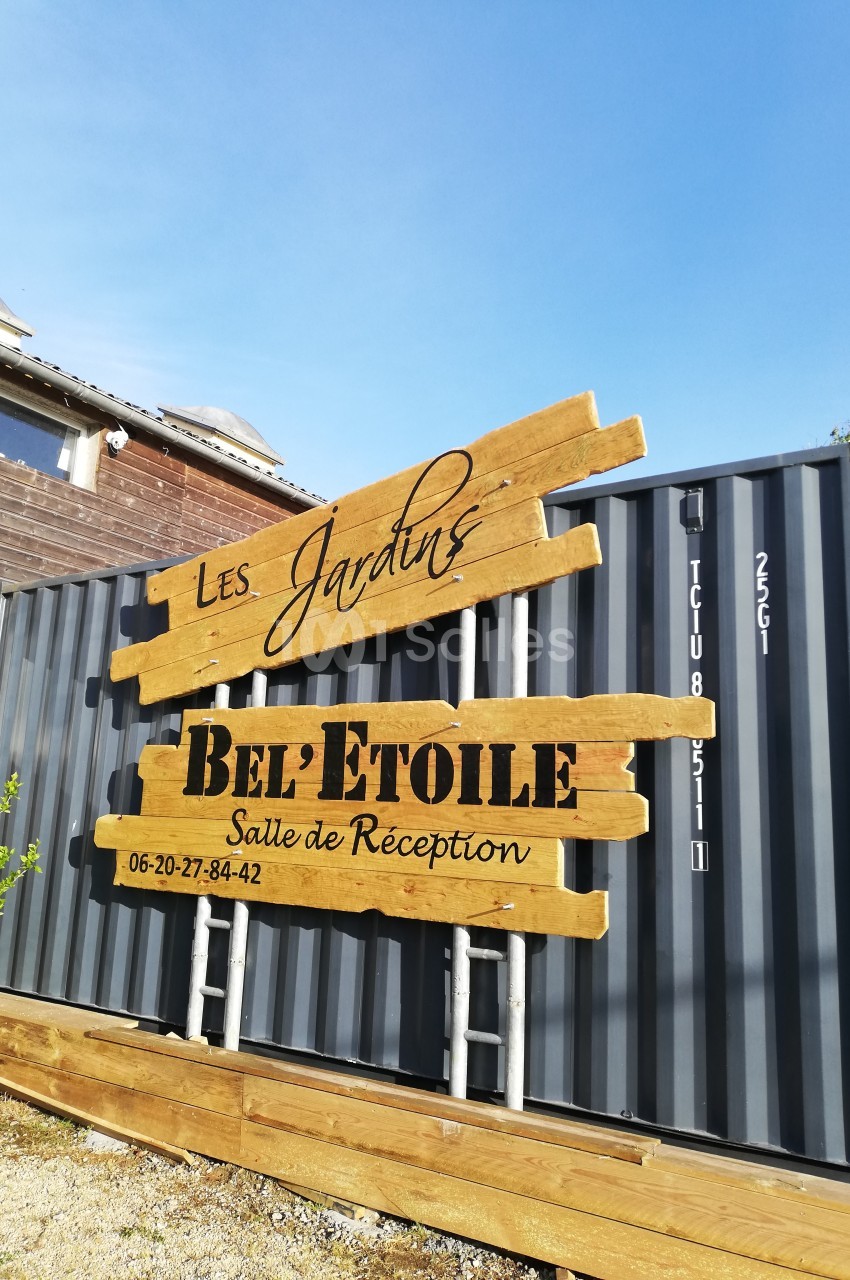 Panneau en bois indiquant ’Les Jardins Bel'Étoile, Salle de Réception’ devant un conteneur gris.