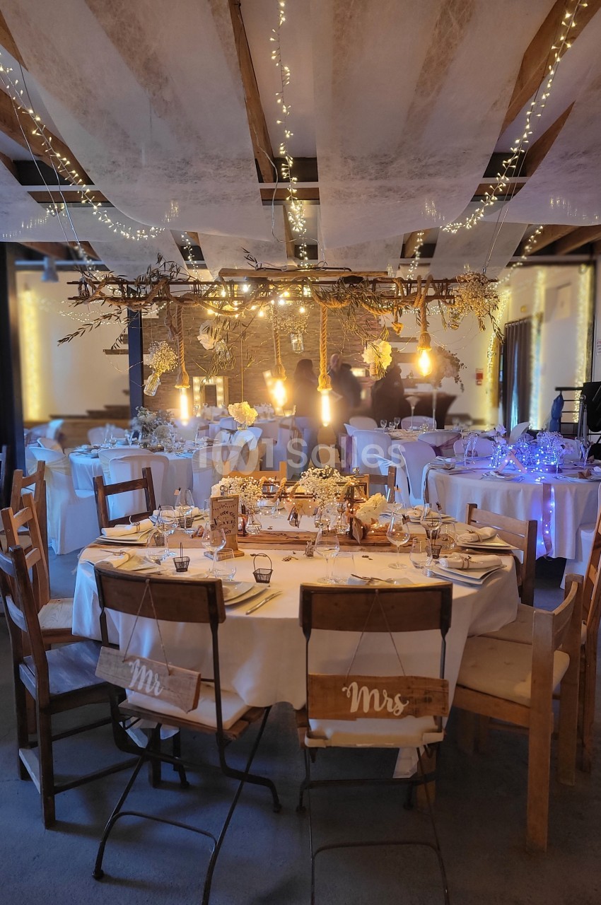 Salle de réception décorée pour un mariage avec guirlandes lumineuses, tables rondes dressées et chaises en bois.