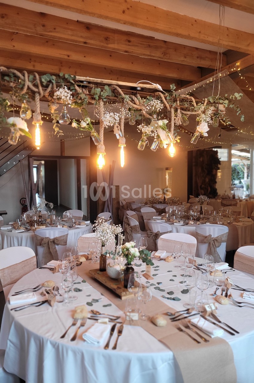 Salle de réception décorée avec des tables rondes, nappes blanches, centres de table fleuris et guirlande lumineuse…
