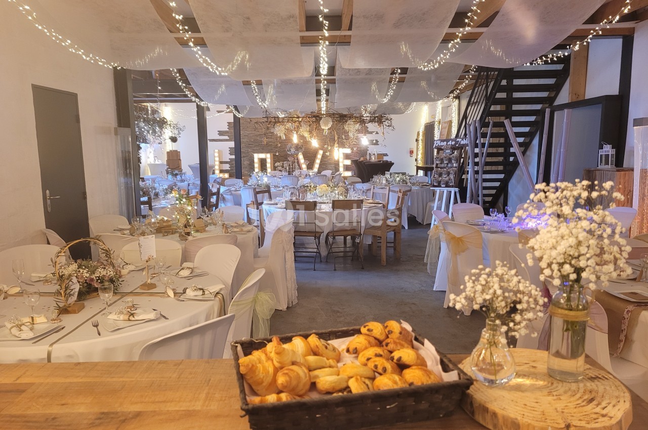 Salle de réception décorée avec des guirlandes lumineuses, des tables dressées et un panier de viennoiseries au premier plan.