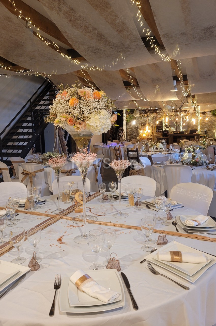 Salle de réception décorée avec des tables rondes élégantes, nappes blanches, fleurs et guirlandes lumineuses suspendues.