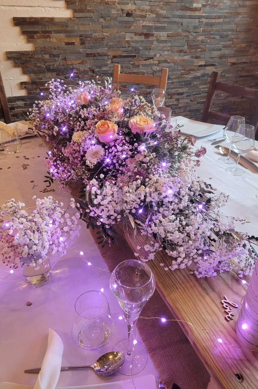 Centre de table floral avec gypsophiles, roses et guirlandes lumineuses violettes sur une table dressée.
