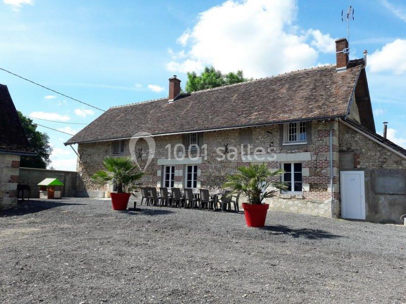 Maison en pierre avec toit en tuiles, grande table extérieure et deux palmiers en pots rouges sur une cour gravillonnée.