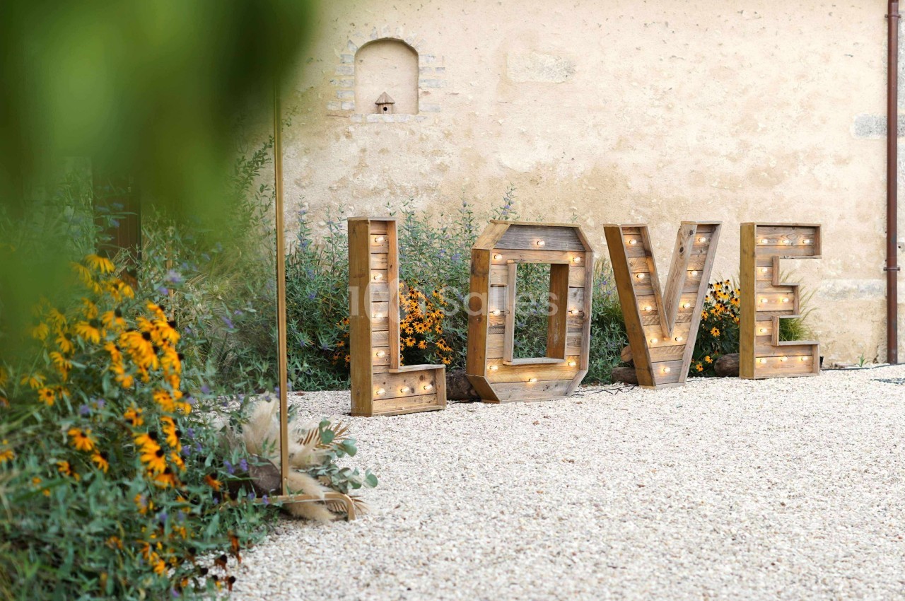 Lettres en bois formant le mot ’LOVE’, éclairées et disposées sur un sol gravillonné devant un mur en pierre.