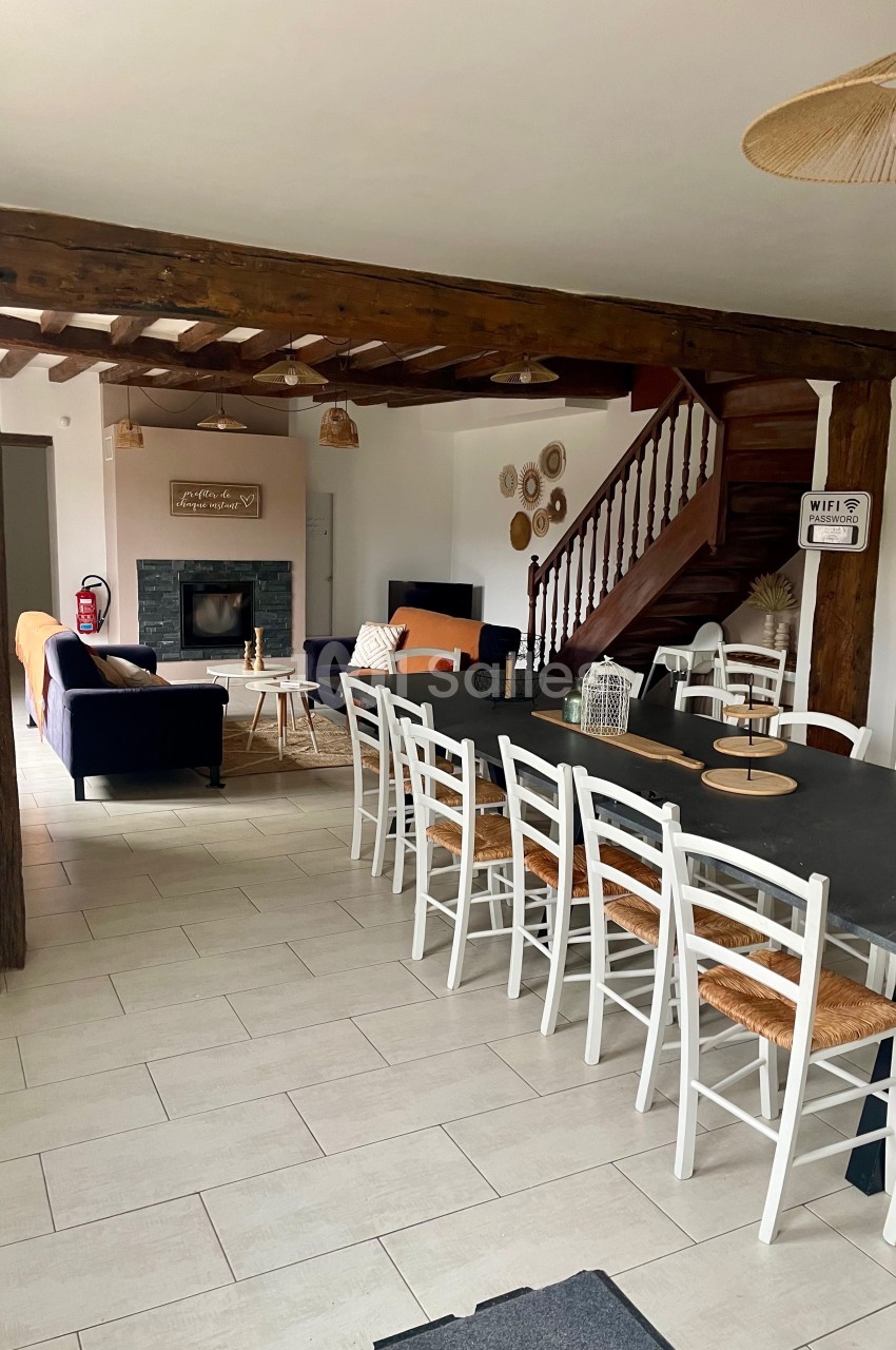 Salle à manger avec une grande table et des chaises blanches, ouverte sur un salon avec cheminée et escalier en bois.