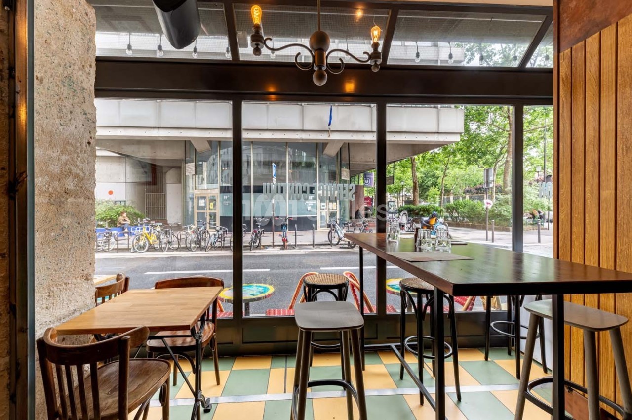 Intérieur d'un café avec des tables en bois, vue sur une rue avec des vélos et un bâtiment moderne à travers une baie vitrée.
