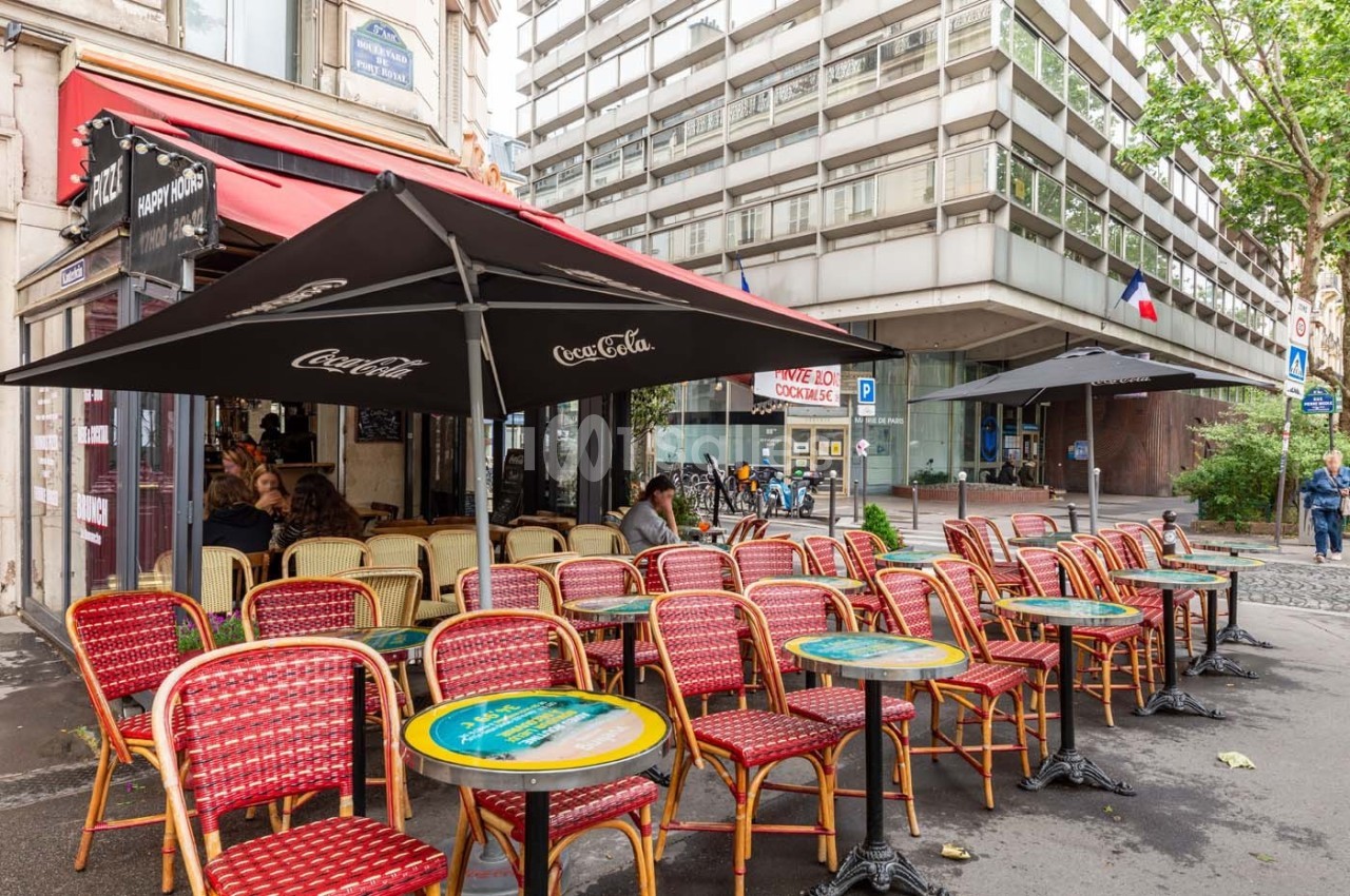 Terrasse de café parisien avec des chaises en osier rouge et des tables rondes, située au bord d'une rue urbaine.