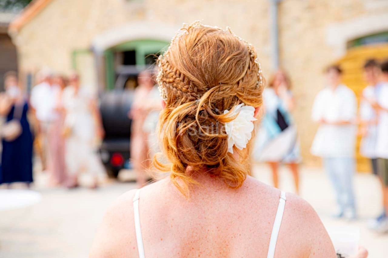 Une femme rousse vue de dos avec une coiffure élaborée et une fleur blanche, lors d'un événement en extérieur.
