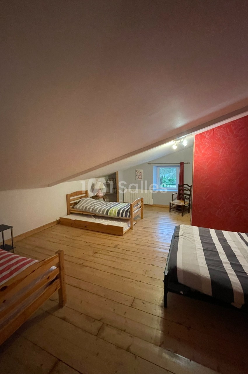 Chambre mansardée avec trois lits simples, parquet en bois, mur rouge décoratif et fenêtre laissant entrer la lumière…