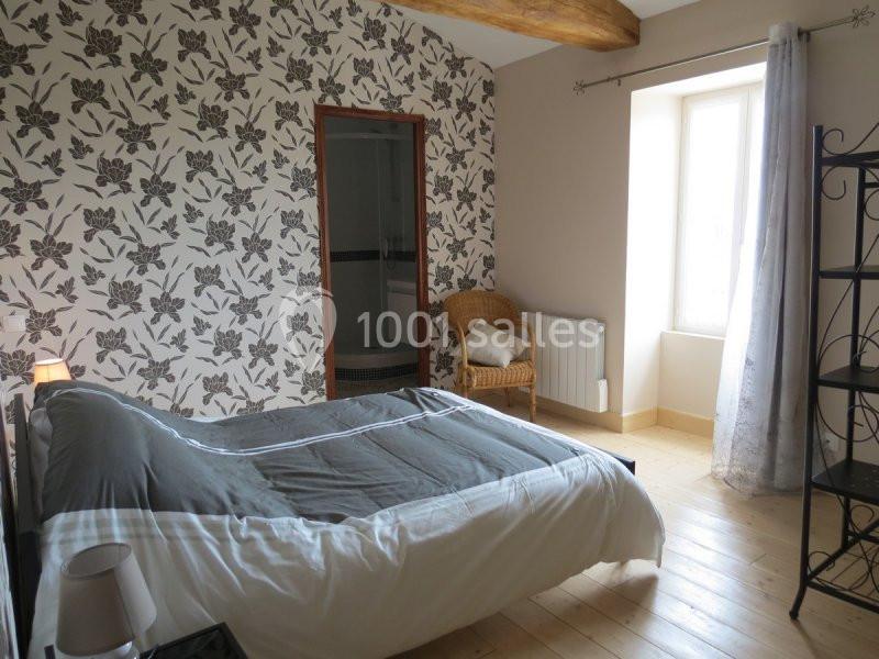 Chambre lumineuse avec lit double, mur à motif floral, parquet en bois clair et fauteuil en osier près d'une fenêtre.